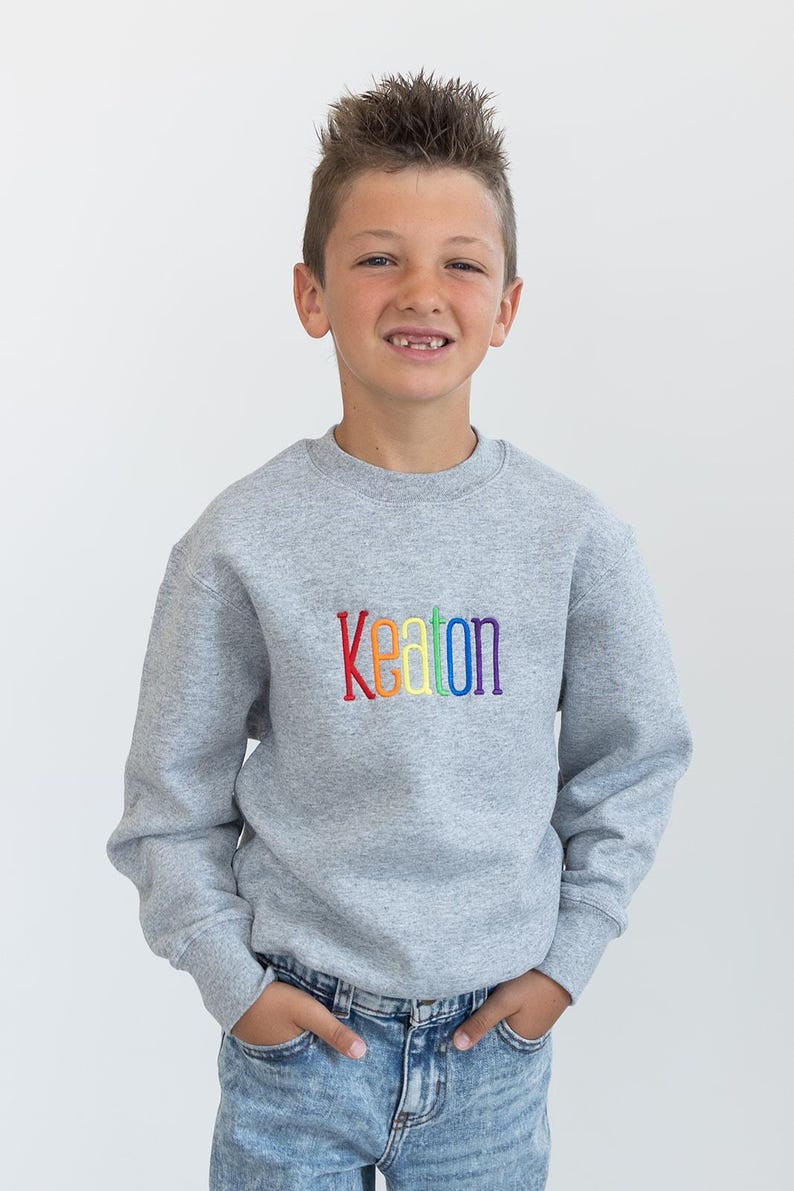 Puede incluir: Una sudadera gris con el nombre "Keaton" bordado en arco&iacute;ris en la parte delantera. La sudadera la lleva puesta un joven que lleva unos vaqueros azules.
