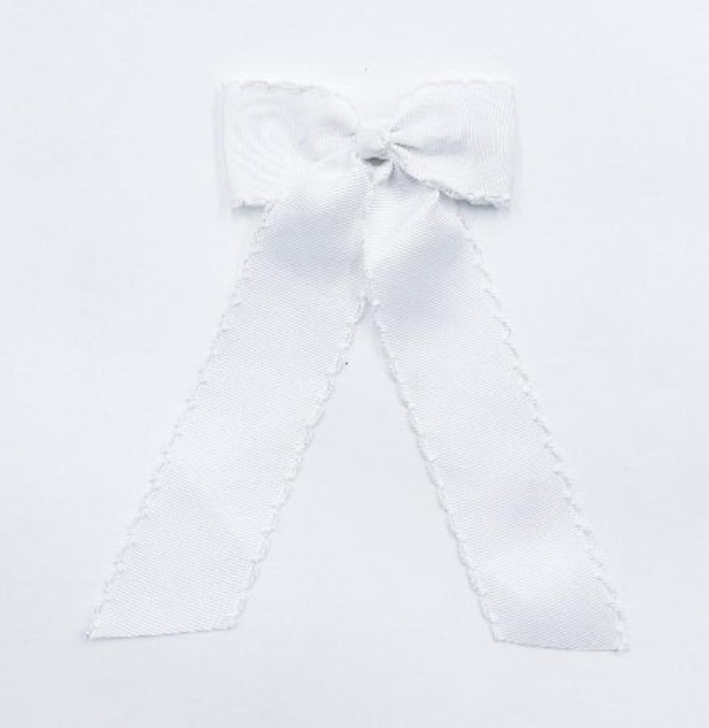May include: A solid white fabric bow featuring a textured grosgrain ribbon construction with a central knot and two long, downward-hanging tails. The edges of the ribbon are finished with a decorative scalloped trim along the entire length.