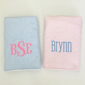 May include: Two baby blankets, one blue with white stripes and the monogram "BSE" in pink, and one pink with white stripes and the name "Brynn" in blue.