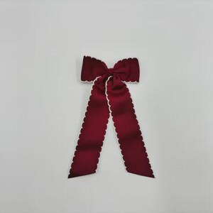 May include: A deep burgundy hair bow featuring a structured knot at the centre and two long, flowing tails. The edges of the fabric are finished with a distinct white scalloped trim that outlines the entire shape of the bow and its long tails.