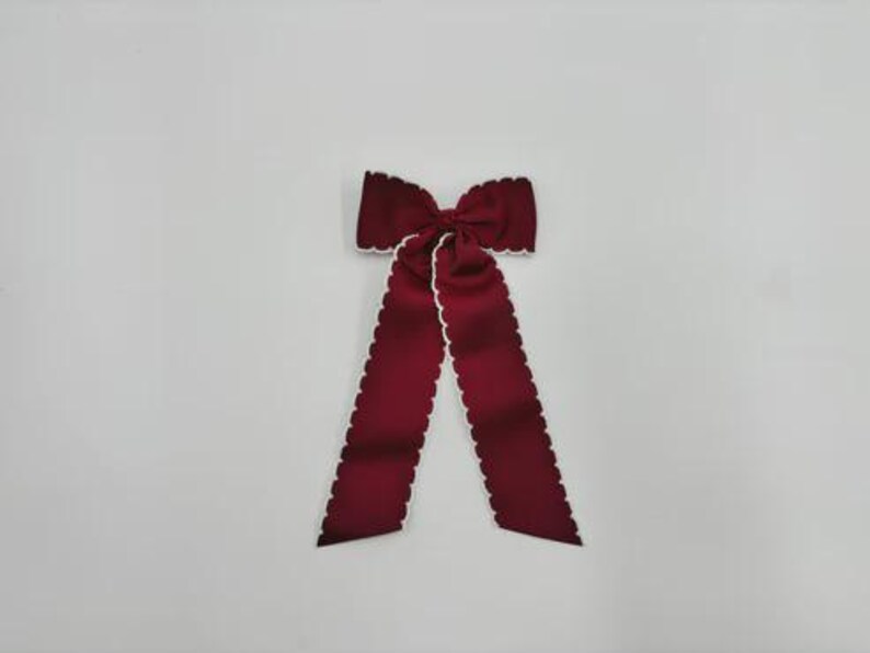May include: A deep burgundy hair bow featuring a structured knot at the centre and two long, flowing tails. The edges of the fabric are finished with a distinct white scalloped trim that outlines the entire shape of the bow and its long tails.