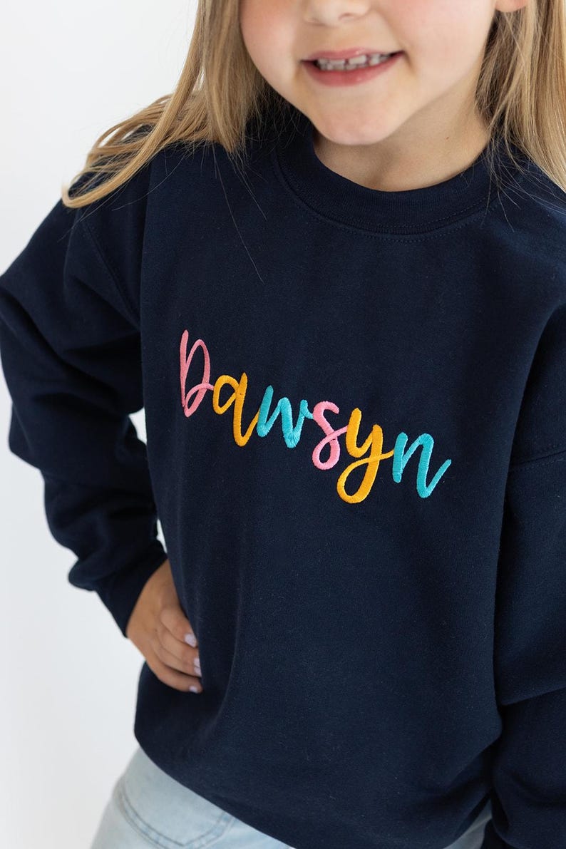 Puede incluir: Una sudadera azul marino con el nombre "Dawson" bordado en letras de colores. Las letras son rosas, amarillas y azules.
