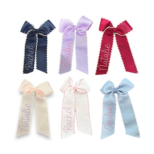 May include: Six hair bows in navy, lavender, maroon, cream, white, and light blue. Each ribbon features scalloped edges and embroidered names including Rachel, Emilia, and Natalie in cursive script. The bows are made of grosgrain fabric.