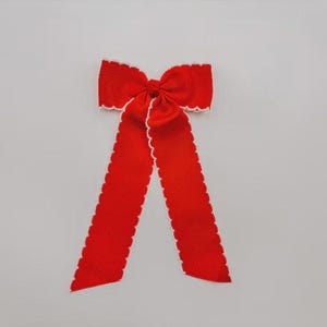 May include: A bright red fabric bow featuring a textured grosgrain ribbon construction. The bow has two long, hanging tails and is finished with a delicate, scalloped white trim along all of its edges, creating a distinct contrast against the red.