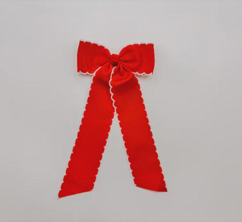 May include: A bright red fabric bow featuring a textured grosgrain ribbon construction. The bow has two long, hanging tails and is finished with a delicate, scalloped white trim along all of its edges, creating a distinct contrast against the red.