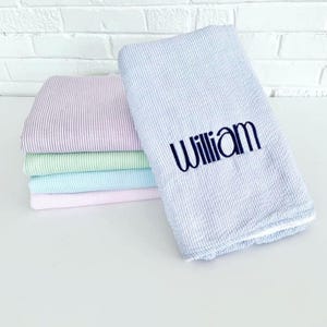 May include: A stack of personalized baby blankets in various pastel colors, including lavender, light green, and light blue. The top blanket is light blue with white stripes and the name "William" embroidered in navy blue.