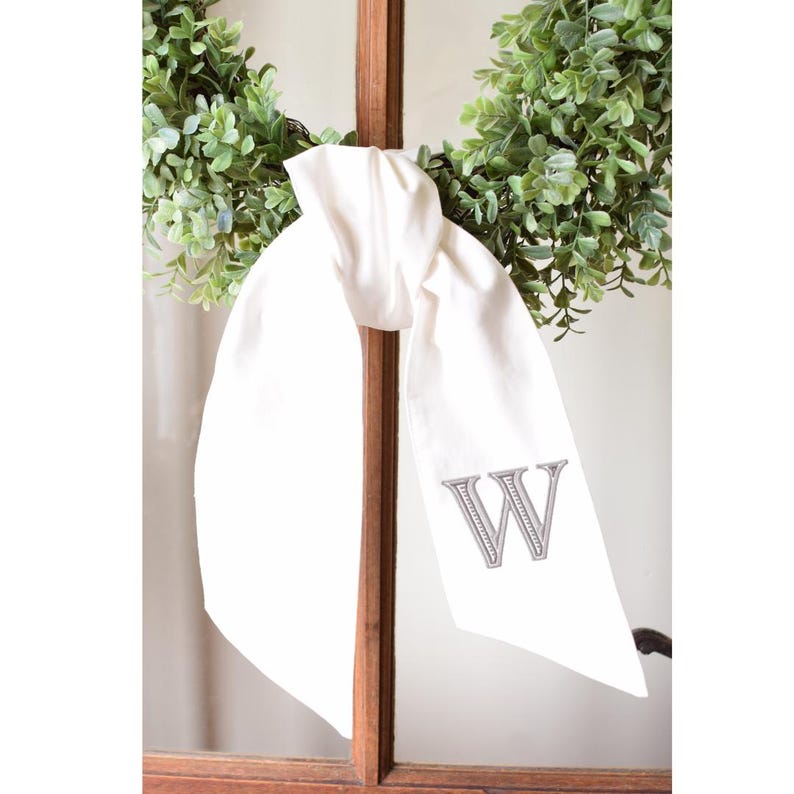 Op de afbeelding: Een wit stoffen lint geknoopt in een strik, voorzien van een grijze geborduurde letter W op een van de hangende uiteinden. Het lint is geplaatst voor een groene bladerkrans en een bruine houten frameconstructie voor decoratieve doeleinden.