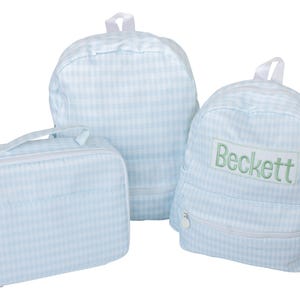 May include: A set of two light blue and white gingham backpacks and one matching rectangular lunch bag. One backpack features a rectangular patch with the name Beckett embroidered in green thread. Each item has white zippers and a top handle.