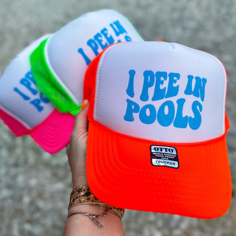 May include: A neon orange trucker hat with a white front panel and a blue 'I PEE IN POOLS' graphic. The hat has a black mesh back and a silver 'OTTO WORLD FAMOUS FOAM FRONT OTTO TRUCKER' label.
