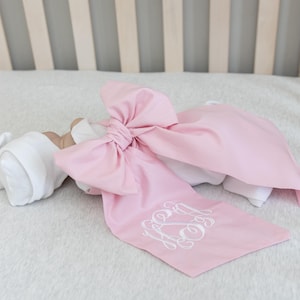 May include: A light pink baby bow with a white monogram is draped over a white blanket. The bow is large and tied in the center. A white cap is visible on the left side of the image. The background includes a crib and a soft, white blanket.