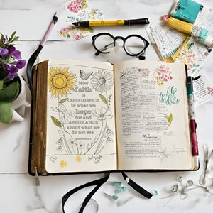May include: An open Bible with a black cover and gold accents. The pages are decorated with hand-drawn flowers and a quote about faith, hope, and assurance. The quote reads "Now faith is confidence in what we hope for and assurance about what we do not see. Hebrews 11:1."