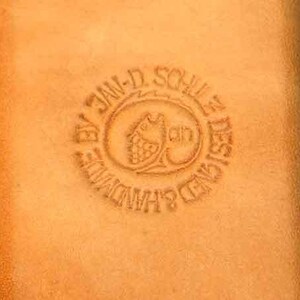 May include: A close-up of a brown leather wallet with a circular embossed stamp that reads "By Jan-D. Schulle Designed Handmade".