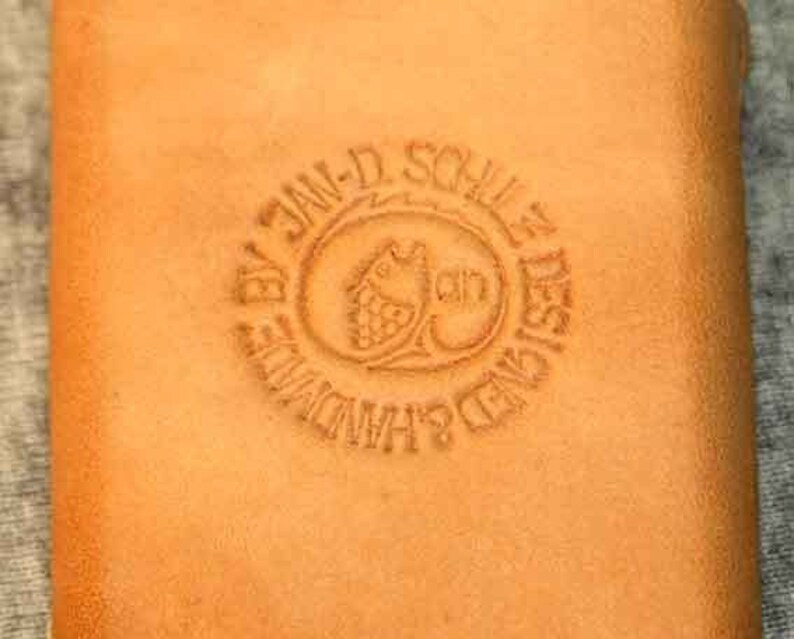 May include: A close-up of a brown leather wallet with a circular embossed stamp that reads "By Jan-D. Schulle Designed Handmade".