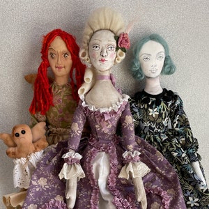 May include: Three handmade fabric dolls with unique designs. The central doll wears a purple floral gown with a ruffled neckline and a powdered wig. The other dolls have red and blue hair, and wear floral dresses. One doll holds a small, tan, stuffed creature.