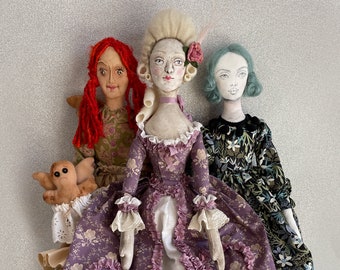 May include: Three handmade fabric dolls with unique designs. The central doll wears a purple floral gown with a ruffled neckline and a powdered wig. The other dolls have red and blue hair, and wear floral dresses. One doll holds a small, tan, stuffed creature.