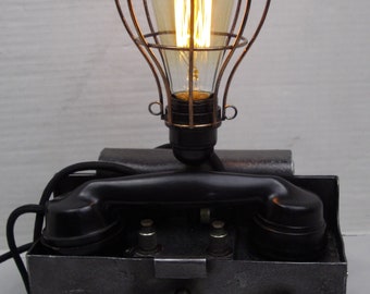 Könnte beinhalten: Eine Schreibtischlampe im Vintage-Stil, hergestellt aus einem umgebauten Drehwahltelefon. Die Lampe verfügt über ein schwarzes Drehwahltelefon mit einem schwarzen Hörer und eine Käfig-Glühbirne mit warmweißem Licht.