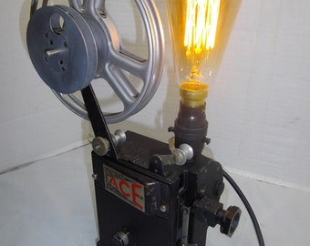 Könnte beinhalten: Eine antike schwarze Filmprojektorlampe mit einer großen silbernen Filmrolle und einer klaren Glühbirne mit warmem gelbem Licht. Der Projektor ist mit "PATENTED ACE SPECIAL" beschriftet.