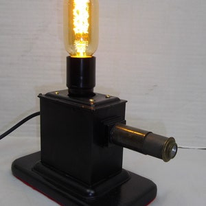 May include: A black metal table lamp with a brass telescope-like arm and a vintage-style light bulb. The lamp has a black base with a red felt bottom.