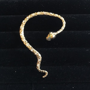 May include: Gold-toned ear cuff in the shape of a snake. The snake's body curves along the ear, adorned with small, clear crystals. The snake's head is detailed with green and gold accents, creating a striking piece of jewelry.