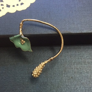 May include: An ear cuff with a calla lily design. The cuff has a curved, gold-toned wire. The lily features a teal enamel leaf with crystal accents and a stem covered in crystals. The cuff is designed to be worn on the ear.