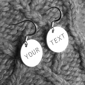 May include: Silver-coloured earrings featuring circular pendants. One pendant is engraved with the word "YOUR", the other with "TEXT". The earrings hang against a grey, knitted fabric background.