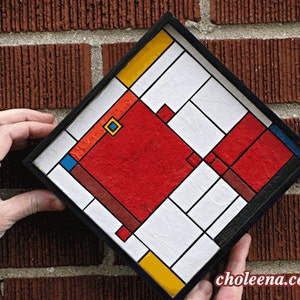 May include: A framed geometric abstract art piece with a red, yellow, blue, and white color scheme. The artwork is made of paper squares and is framed in black.