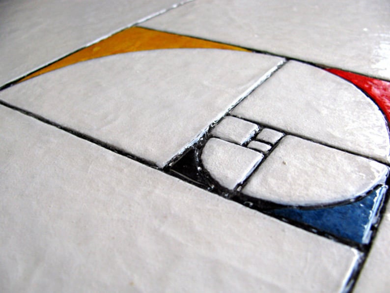 May include: Close-up of a geometric design featuring a Fibonacci sequence spiral. The design is composed of white, square tiles with black grout, and segments of yellow, red, and blue. The overall aesthetic is modern and artistic.