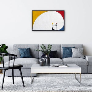 May include: A framed abstract art piece featuring the golden ratio. The artwork has a white background with a yellow, red, and blue curved design. The painting is displayed above a gray sofa with decorative pillows, a white coffee table, and a black chair.