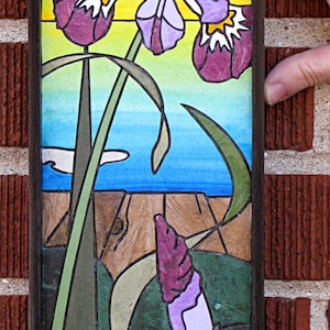 May include: A framed stained glass artwork featuring purple iris flowers with green stems and leaves set against a blue and yellow background. The artwork is titled "Art by Choleena" and includes the website "choleena.com".