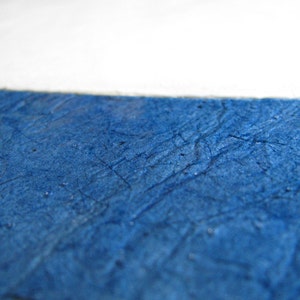 May include: Close-up of textured, deep blue paper with a white section at the top. The paper has a crinkled appearance with visible lines and subtle variations in color, creating a tactile and artistic effect. The image suggests a focus on texture and color.