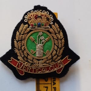 Puede incluir: Parche bordado con fondo negro, corona de laurel dorada y centro verde con dise&ntilde;o floral. El parche presenta una corona en la parte superior y las palabras "VERTICAL CLUB" en una pancarta roja.