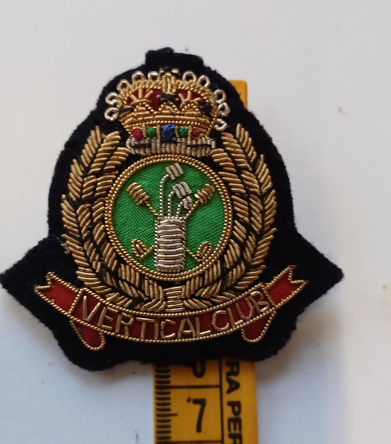 Puede incluir: Parche bordado con fondo negro, corona de laurel dorada y centro verde con dise&ntilde;o floral. El parche presenta una corona en la parte superior y las palabras "VERTICAL CLUB" en una pancarta roja.