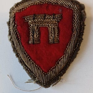 Puede incluir: Un parche vintage en forma de escudo con un centro rojo y un dise&ntilde;o bordado en color dorado. El dise&ntilde;o parece ser un elemento arquitect&oacute;nico estilizado. El parche est&aacute; bordeado con hilo dorado y marr&oacute;n.