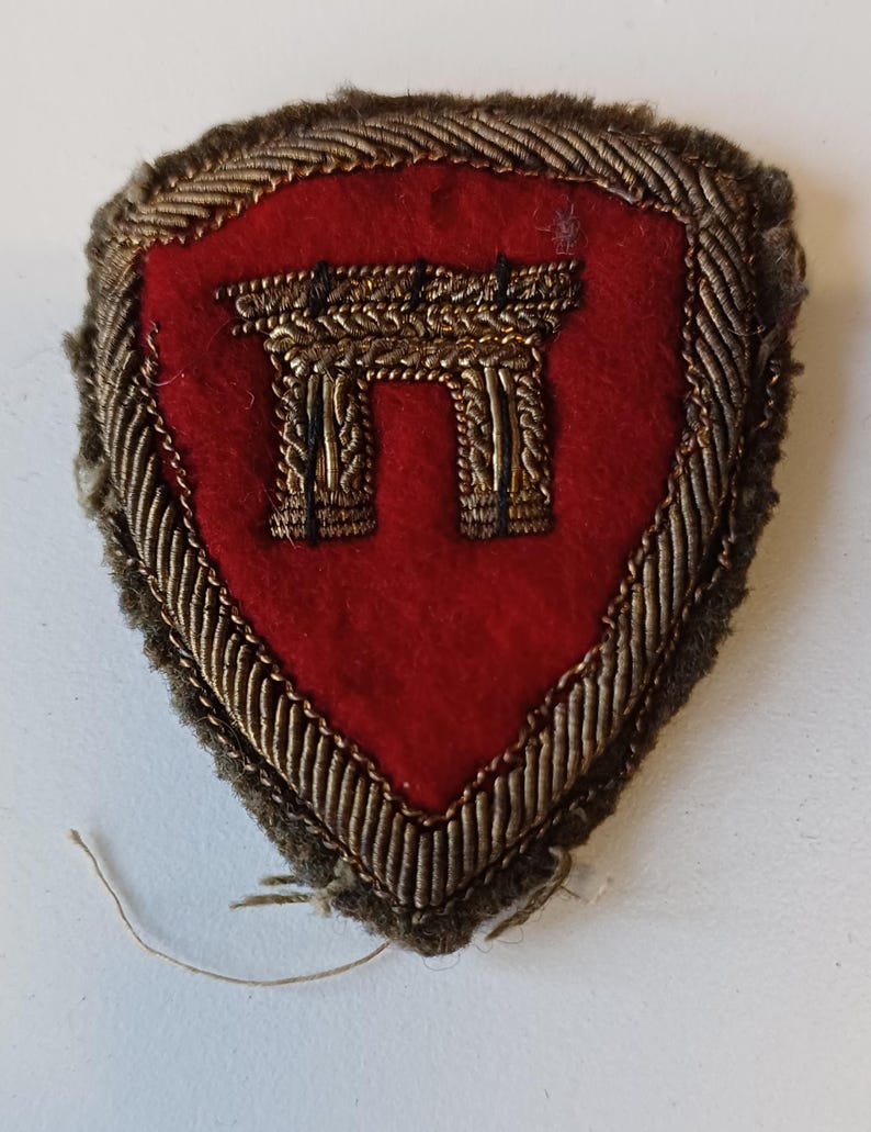 Puede incluir: Un parche vintage en forma de escudo con un centro rojo y un dise&ntilde;o bordado en color dorado. El dise&ntilde;o parece ser un elemento arquitect&oacute;nico estilizado. El parche est&aacute; bordeado con hilo dorado y marr&oacute;n.