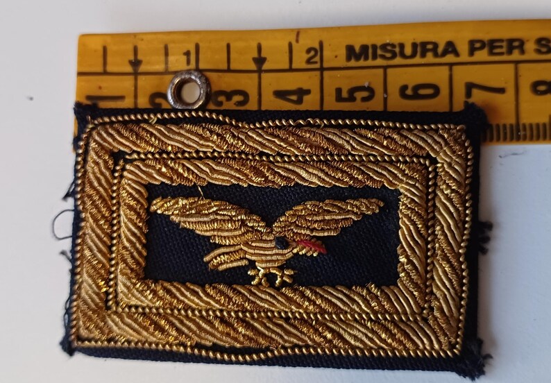 Puede incluir: Parche rectangular con un emblema de &aacute;guila dorada sobre fondo azul marino, enmarcado por un ribete trenzado dorado. El &aacute;guila tiene un pico rojo y est&aacute; centrada en el parche. Un anillo met&aacute;lico est&aacute; fijado en la parte superior.