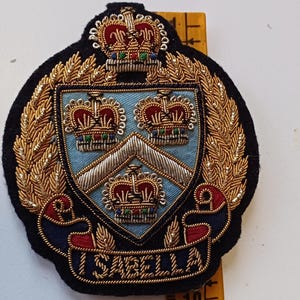 Puede incluir: Un escudo bordado con un dise&ntilde;o de escudo, con tres coronas y el nombre "ISABELLA" en una pancarta. El escudo est&aacute; rodeado de adornos dorados y sobre un fondo negro.