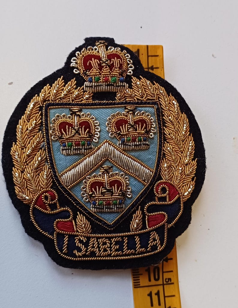 Puede incluir: Un escudo bordado con un dise&ntilde;o de escudo, con tres coronas y el nombre "ISABELLA" en una pancarta. El escudo est&aacute; rodeado de adornos dorados y sobre un fondo negro.