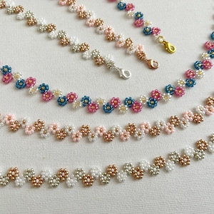 May include: A selection of beaded necklaces and bracelets with a floral pattern. The jewellery features small, colourful beads in pink, blue, white, and gold. The pieces are displayed on a white surface.