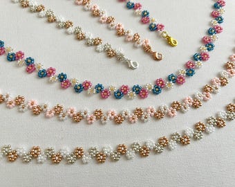 Könnte beinhalten: Mehrere Halsketten und Armbänder aus Perlen mit floralem Design. Der Schmuck besteht aus kleinen, farbigen Perlen in Rosa, Blau, Weiß und Gold. Die Stücke sind auf einer weißen Oberfläche angeordnet.