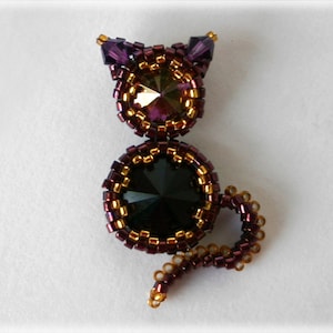 Könnte beinhalten: Eine gefertigte Katzenbrosche mit einem schwarzen Kristall in der Mitte, umgeben von goldenen und violetten Perlen.