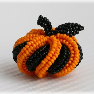 Op de afbeelding: Een kleine, geperde pompoen met een zwarte steel en bladeren. De pompoen is gemaakt van oranje en zwarte kralen, waardoor een gestreept patroon ontstaat.