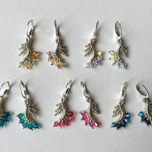 Può includere: Sei paia di orecchini pendenti con elementi metallici color argento e accenti di cristalli colorati. Gli orecchini presentano un design a forma di foglia con vari colori di cristallo, tra cui giallo, arancione, viola, verde acqua e rosa. Sono esposti su una superficie bianca.