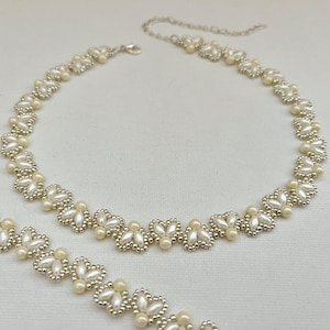 May include: A delicate white and silver beaded necklace with a floral design. The necklace is made of small, round beads that are strung together in a pattern of flowers. The necklace is perfect for adding a touch of elegance to any outfit.