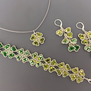 Op de afbeelding: Een set groene en zilveren kralen sieraden met een ketting, armband en oorbellen. De sieraden zijn gemaakt van kleine, gefacetteerde kralen die in een klaverbladvorm zijn gerangschikt. De ketting heeft een enkele klaverblad hanger, terwijl de armband en oorbellen meerdere klaverbladeren hebben.
