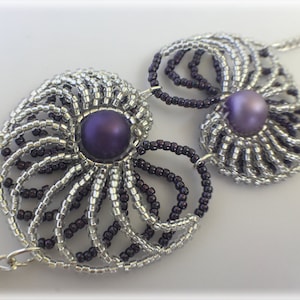 May include: A silver and purple beaded necklace with two large, round, purple beads in the center. The beads are surrounded by silver and purple beads in a spiral pattern.