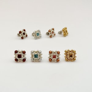 Può includere: Un set di dieci orecchini a forma quadrata con pietre centrali colorate. Gli orecchini sono realizzati con piccole perline bianche e presentano una varietà di colori, tra cui rosso, blu e oro.