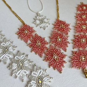 May include: A collection of beaded jewelry featuring floral designs. The set includes a silver and white beaded bracelet, a silver pendant necklace, and necklaces and a bracelet in coral and gold. The jewelry is displayed on a white surface.