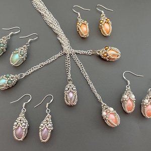 Puede incluir: Un conjunto de joyas de cuentas que incluye un collar y pendientes. El collar tiene una cadena plateada y un colgante con una cuenta en forma de lágrima de color morado claro. Los pendientes tienen ganchos plateados y cuentas en forma de lágrima en tonos verdes, rosas y melocotón.