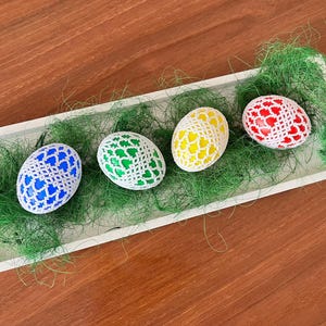 May include: Four Easter eggs decorated with white crocheted lace. The eggs are coloured blue, green, yellow, and red.