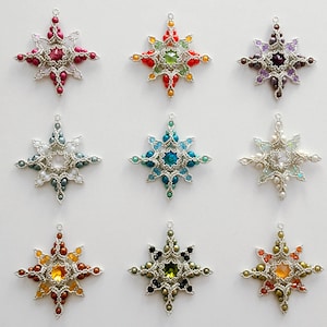 Może przedstawiać: Dziewięć koralikowych ozdób w kształcie gwiazd w różnych kolorach, w tym czerwonym, zielonym, niebieskim i fioletowym. Każda gwiazda ma centralny klejnot i jest otoczona mniejszymi koralikami, wszystkie przymocowane do srebrnej ramy. Idealne do dekoracji świątecznych.